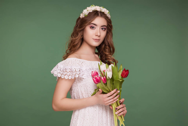 A beautiful young woman stands on a purple background, wearing a white dress and a wreath of flowers on her head, and a bouquet of tulips. International Women's Day, March 8. - Foto, imagen