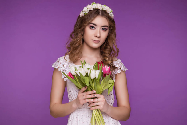 A beautiful young woman stands on a purple background, wearing a white dress and a wreath of flowers on her head, and a bouquet of tulips. International Women's Day, March 8. - Valokuva, kuva