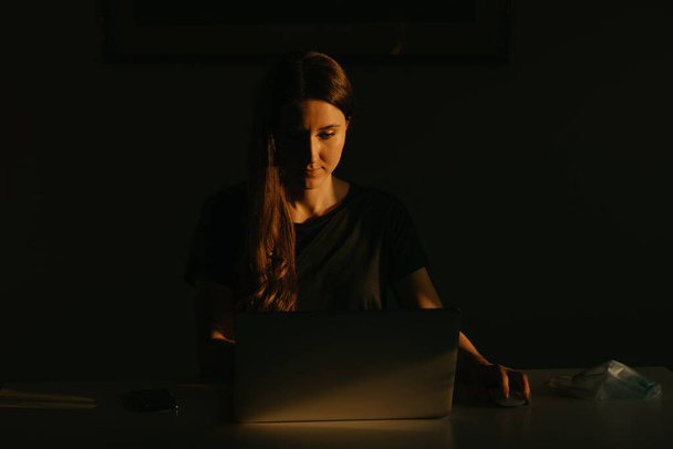 A caucasian woman working remotely on her laptop during the quarantine to avoid the spread coronavirus. A businesswoman working from home in the evening during the pandemic of COVID-19.  - Photo, Image