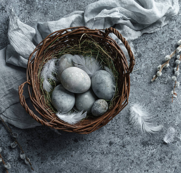 Huevos de Pascua en canasta de mimbre con plumas y flores de primavera sobre fondo azul. Felices vacaciones de Pascua, vista superior, acostado plano
 - Foto, imagen