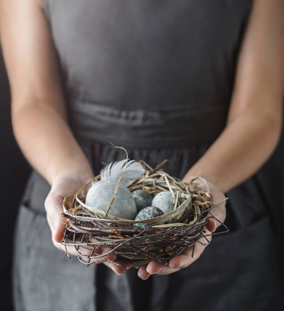 Жінка тримає гніздо з мармуровими яйцями Пасхи з пір'ям на темному тлі зі світлом. З Великоднім святом, з вибірковим фокусом. - Фото, зображення