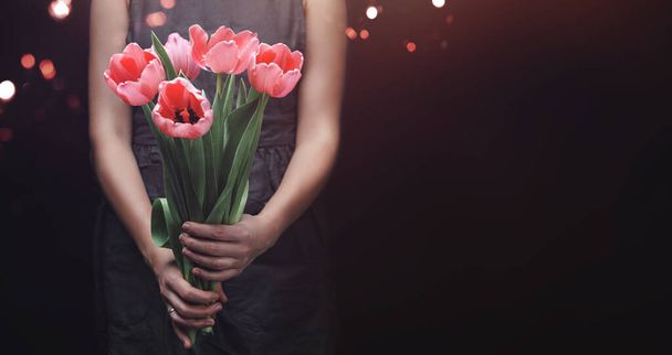 女性の手は、光、ボケと暗い背景にピンクのチューリップの花を保持。ハッピーイースター休暇,バレンタインデー,女性の日,選択的フォーカス - 写真・画像