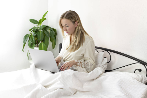 Jonge mooie Europese blonde vrouw werkt en studeert thuis in bed met laptop en smartphone. ze is in lichte, comfortabele huiskledij. - Foto, afbeelding