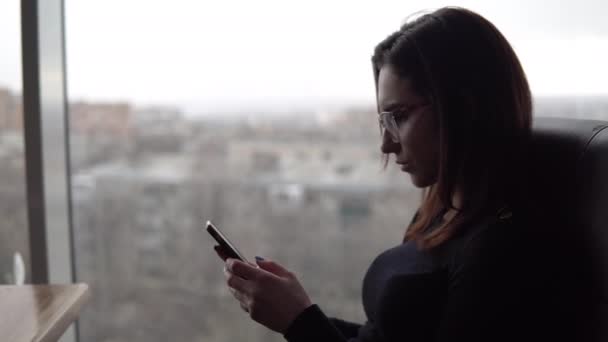 A young woman with a phone sits at a table. A girl sits in a cafe by the panoramic window with a smartphone in her hands. View from the window. - Filmati, video
