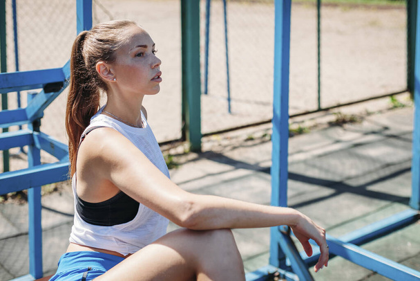 Athletic young blond woman sits and rests on the sports field during her vacation from workouts. - Valokuva, kuva