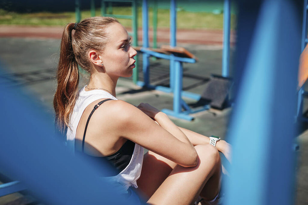 Αθλητική νεαρή ξανθιά γυναίκα κάθεται και ξεκουράζεται στο γήπεδο κατά τη διάρκεια των διακοπών της από προπονήσεις. - Φωτογραφία, εικόνα