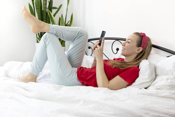 joven mujer sonriente con el pelo rubio en una camiseta roja y pantalones cómodos se encuentra en una cama blanca con un teléfono inteligente. trabaja, estudia desde casa, escribe y lee mensajes en el móvil
, - Foto, imagen