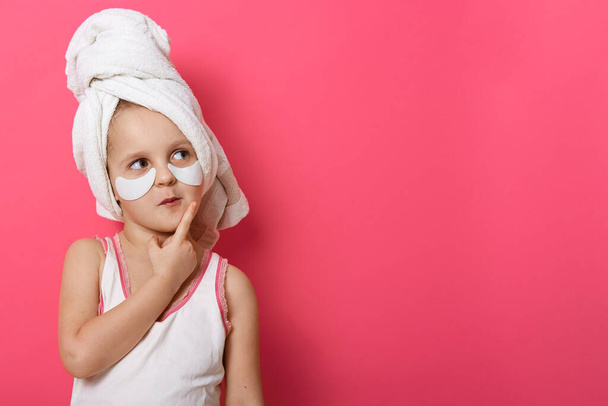 Image of little girl with white towel on her head, having patches under her eyes, thinking about something nice, keeping finger near mouth. Copy space for your advertisment or promotional text. - Foto, imagen