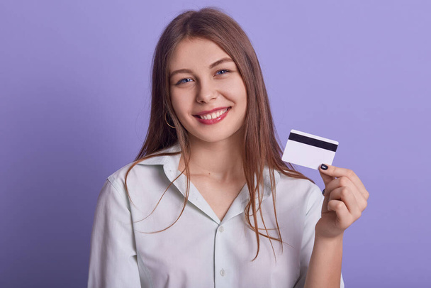 Genç ve güzel bir iş kadınının portresi elinde kredi kartıyla eflatun arkaplanından izole edilmiş poz veriyor doğrudan kameraya bakıyor internetten alışveriş yapan beyaz bluz giyen bir kadın.. - Fotoğraf, Görsel