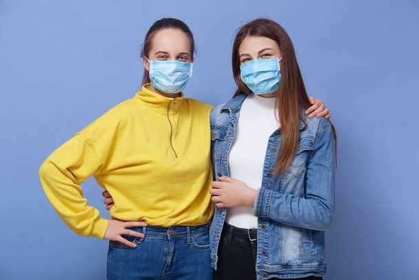 Close up portrait of two young females wearing protective disposable medical masks on faces for corona virus spread prevention, lady dresses yellow shirt hugs her friend, looking at camera. Covid 19. - 写真・画像