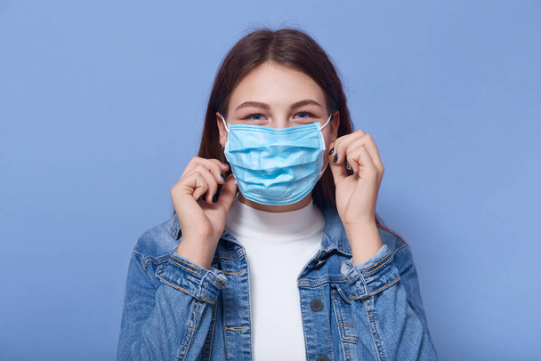 Indoor shot of woman wearing disposable protective face mask. Air pollution, industrial emissions or virus epidemic concept, female wearing denim jacket and white shirt stands over lilac studio wall. - Фото, изображение