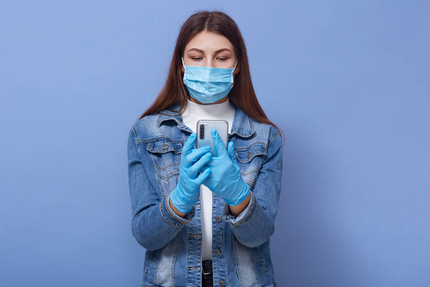 Close up portrait of young woman wearing medical face mask and blue gloves typing on mobile phone isolated over lilac background, girl wearing white shirtand denim jacket. Coronavirus concept. - Foto, Imagem