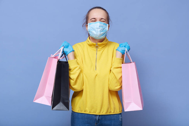 Studio shot of young girl dresses medical mask, protective gloves and casual outfit clutching shopping bags in her hands, keeps eyes closed, looks happy, protects herself while doing shopping. - 写真・画像