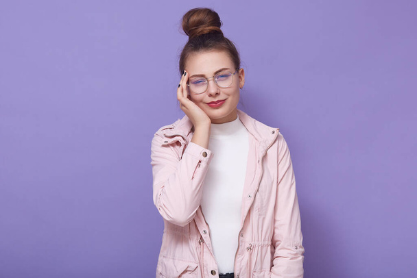 Horizontal shot of unhappy woman wearing glasses, jacket and white shirt, looks tired, massaging touching temple, exhausted girl suffering from headache, posing isolated over lilac studio background. - Photo, Image