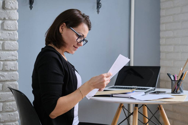 Mature woman sitting at her desk with paper letter document. Smiling female reading good news, envelopes, laptop on the table - Φωτογραφία, εικόνα