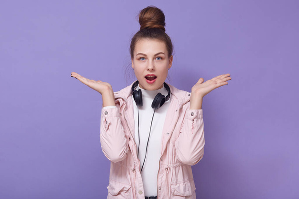 Indoor shot of confused european female with hair bun, spreading hands and standing with opened mouth, having surprised facial expression, posing isolated over lilac wall, has headphones around neck. - Fotoğraf, Görsel