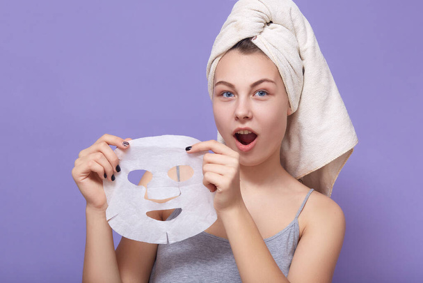 Portrait of astonish beautiful woman with white towel on head and perfect skin, taking off fabric face mask isolated over lilac studio background. Concept of cosmetic and skin care procedures. - Foto, Imagen