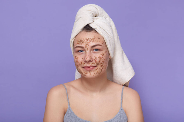 Horizontal shot of teenage girl cleaning her face with coffee scrub, young age female having some difficulties with fece, doing home procedures, peeling her face, wearing white towel and gray t shirt. - Photo, Image