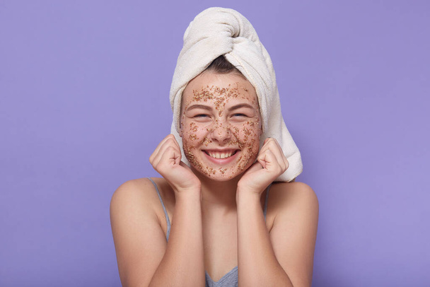 Headshot of smiling happy dark skinned female model has rejuvenation procedure, cleans face with white crystal peeling mask after taking shower, looks younger, isolated over pink background. - Foto, afbeelding