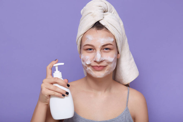 Close up portrait of young attractive girl holding in hands soap dispenser, being ready to remove fasial mask off, posing isolated over lilac background, skin care procedures at home in morning. - Photo, Image