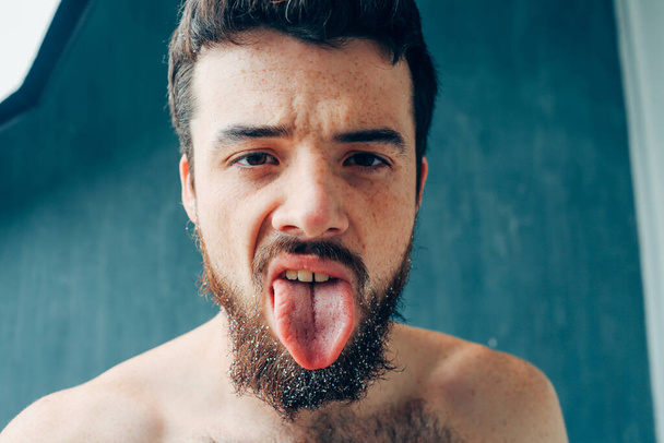 Young man isolated over blue background. Weird guy show long tongue. Keep it outside of mouth. Show on camera its lenghts. Emotional man. - Foto, imagen