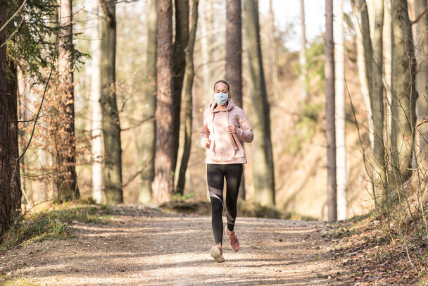 Das Coronavirus oder Covid-19 breitet sich auf der ganzen Welt aus. Porträt einer kaukasischen sportlichen Frau, die beim Laufen in der Natur eine medizinische Schutzmaske trägt. - Foto, Bild