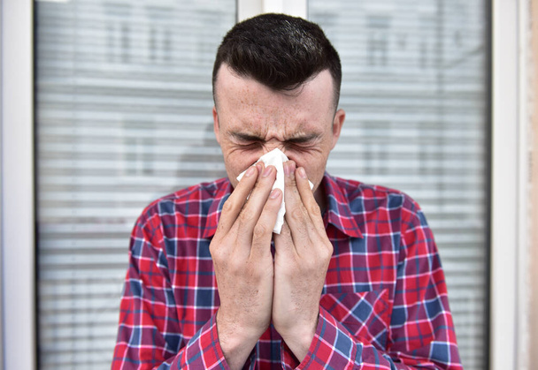 シャツを着た若い男が白いナプキンにくしゃみをする。病気になった風邪やコロナウイルスをキャッチします。インフルエンザと頭痛だ。症状が悪い。正面窓の外に立つ. - 写真・画像