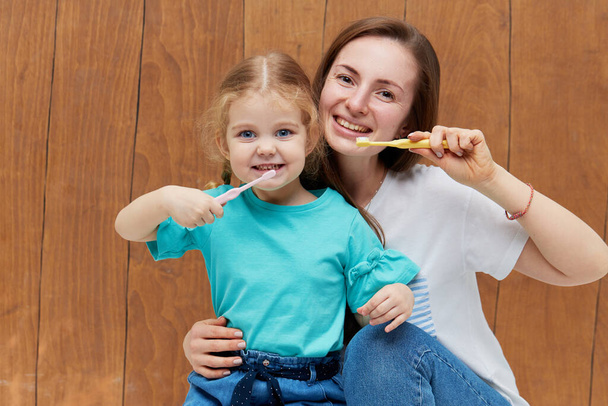 Oral hygiene. Little child girl with mother brushing her teeth with a toothbrush and looking at the camera  on wooden background - Foto, afbeelding