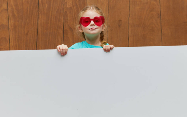 Malé usměvavé dívčí brýle ve tvaru srdce, stojící za bílým prázdným panelem na dřevěném pozadí. Vtipná tvář. Nahlédnutí zpoza praporu, prázdné místo pro text. - Fotografie, Obrázek
