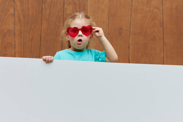 Klein lachend kind meisje hartvormige glazen staan achter een wit blanco paneel tegen houten achtergrond. Grappig gezicht. Achter een banner naar buiten gluren, lege ruimte voor tekst. - Foto, afbeelding