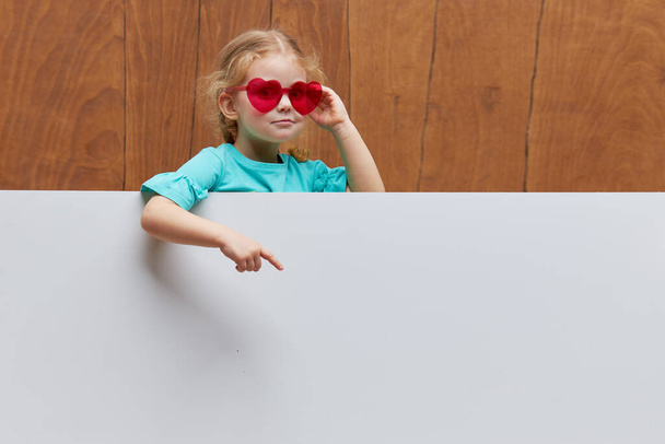 Little smiling child girl heart shaped glasses standing behind a white blank panel against wooden background. Funny face. Peeking out from behind a banner, empty space for text. - Photo, Image