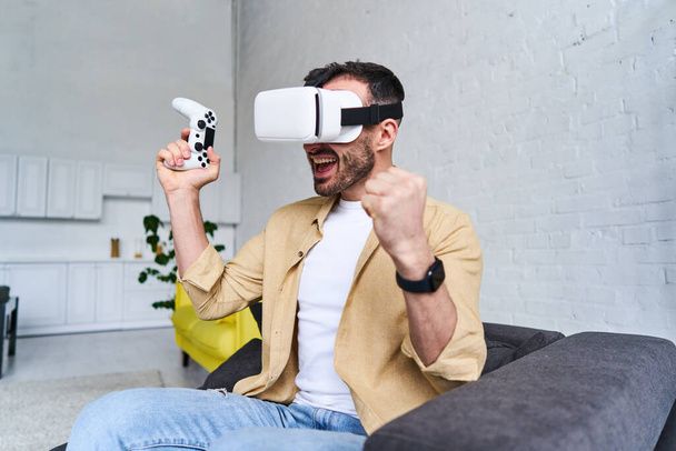 Молодой эмоциональный человек в очках виртуальной реальности играет в видеоигру с джойстиком, удовлетворенностью, счастьем, волнением, радостью победы, победой
 - Фото, изображение