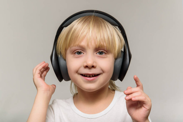 Szőke fiú fülhallgatóban, világos háttérrel. A gyerek zenét hallgat és mosolyog.. - Fotó, kép