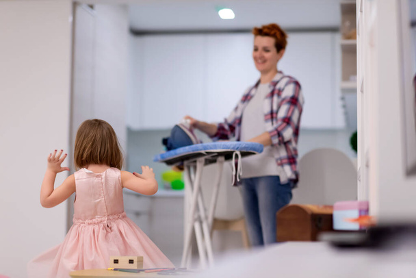 happy family having fun together at home  cute little daughter in a pink dress playing and dancing while young redhead mother ironing clothes behind her - Photo, image