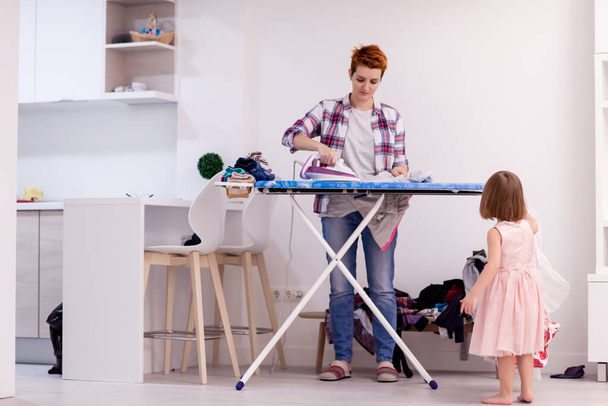happy family having fun together at home  cute little daughter in a pink dress playing and dancing while young redhead mother ironing clothes behind her - Photo, Image