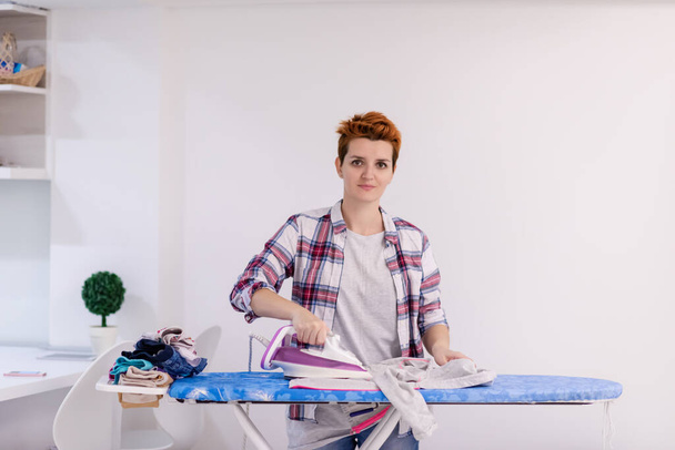 beautiful young red haired housewife in good mood ironing clothes on ironing board in front of a white wall at home - Foto, immagini
