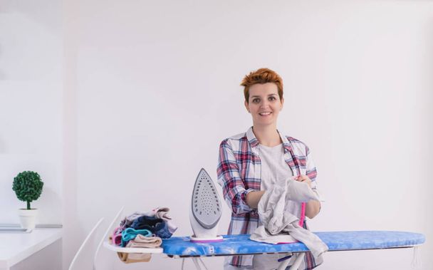 beautiful young red haired housewife in good mood ironing clothes on ironing board in front of a white wall at home - Foto, Imagem