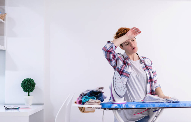 beautiful young red haired housewife in good mood ironing clothes on ironing board in front of a white wall at home - Foto, Imagem
