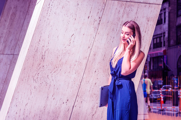 Jeune Américaine d'Europe de l'Est parlant au téléphone, voyageant, travaillant à New York, portant une combinaison bleue, portant un ordinateur portable, sortant de l'immeuble de bureaux. Effet filtré par couleur
 - Photo, image