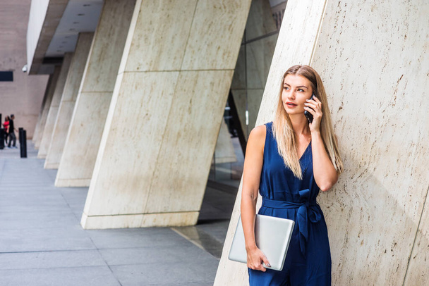 Young Eastern European American Woman with long brown hair, wearing blue sleeveless jumpsuit, holding laptop computer, standing against column outside office in New York City, talking on cell phone - Fotografie, Obrázek