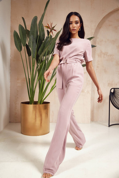 Beautiful sexy brunette woman tanned skin face cosmetic makeup wear pink suit pants for date walk office fashion clothes style collection interior room  sand color safari summer armchair palm boho. - 写真・画像