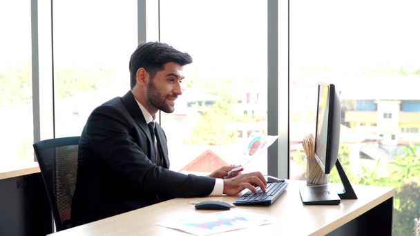 Ευτυχισμένος νέος επαγγελματίας επιχειρηματίας που κάθεται στο γραφείο και εργάζεται και κοιτάζει οθόνες υπολογιστών και έγγραφα στο χώρο εργασίας. Η έννοια της ανάλυσης δεδομένων και της online τεχνολογίας - Φωτογραφία, εικόνα
