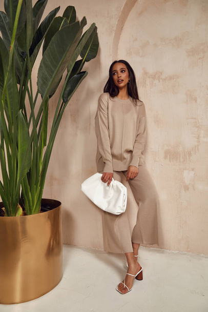 Beautiful sexy brunette woman face cosmetic makeup tanned skin wear fashion clothes style collection beige knitted suit sandals accessory  bag interior sand safari journey summer wall flowerpot palm. - Foto, Bild