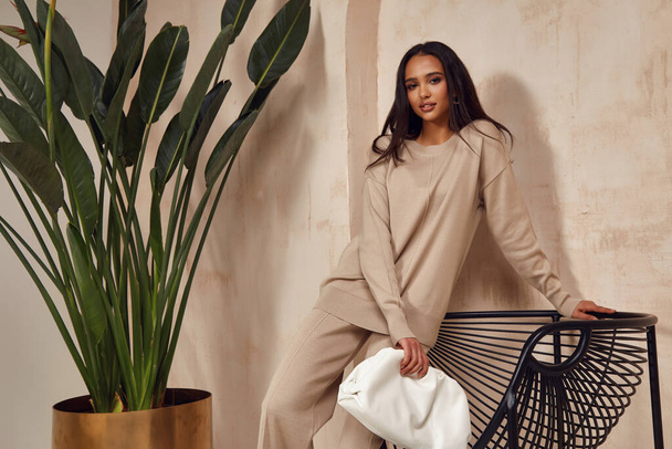 Beautiful sexy brunette woman face cosmetic makeup tanned skin wear fashion clothes style collection beige knitted suit sandals accessory bag interiorfurniture armchair journey summer wall flowerpot palm boho. - 写真・画像