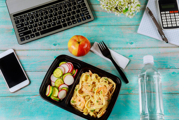 Lunch box container with pasta and shrimps, cucumber and radish on wooden table with laptop. Food delivery concept. - Fotografie, Obrázek