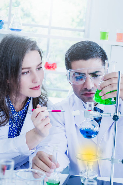 Een Midden-Oosterse mannelijke wetenschapper en Kaukasische mooie vrouwelijke wetenschapper analyseren de groene oplossing in de flits in een wetenschappelijk laboratorium. - Foto, afbeelding