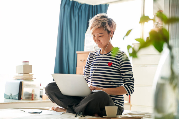 Азиатская женщина сидит на кровати и работает из дома на ноутбуке
 - Фото, изображение