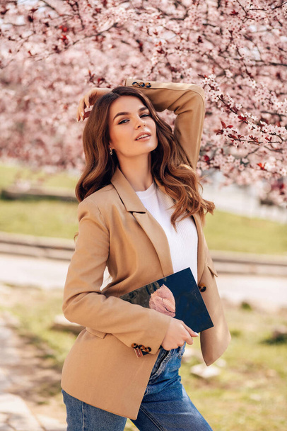 fashion  photo of beautiful woman with dark hair in elegant clothes posing among blossoming peach trees in park - Foto, afbeelding
