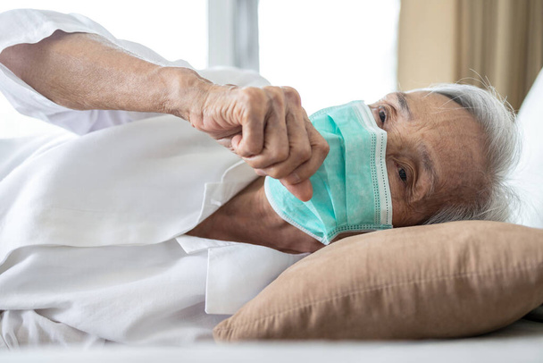 Άρρωστη Ασιάτισσα ηλικιωμένη γυναίκα στο κρεβάτι, φοράει μάσκα προστασίας προσώπου, πυρετό και βήχα, πνευμονία, ηλικιωμένη ασθενής έχει ασθένεια, γρίπη και κρυολόγημα, συμπτώματα του Covid-19, πανδημία, ηλικιωμένοι άνθρωποι μολυσμένοι με τον ιό Coronavirus   - Φωτογραφία, εικόνα
