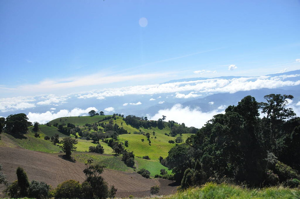 Pendientes agrícolas del volcán Irazu en Costa Rica con suelo fértil y numerosos cultivos en laderas soleadas. El valle está rodeado de nubes suspendidas bajo el cono volcánico
. - Foto, Imagen